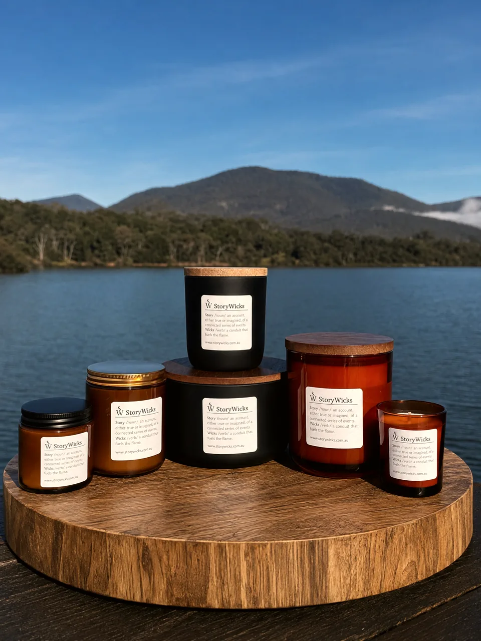 Six StoryWicks candles in amber glass and matte black vessels with timber and gold lids, grouped on a round timber board beside a lake with forested hills behind.