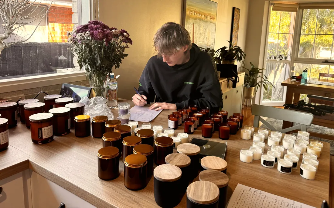 A home workbench covered with dozens of amber glass jars, matte black vessels and small votives, with a maker sitting at the centre writing labels.